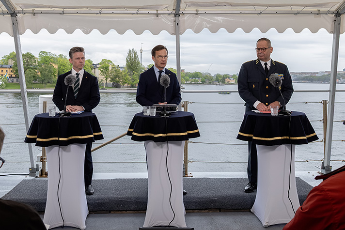 Minister for Defence Pål Jonson, Prime Minister Ulf Kristersson and Chief of Defence Michael Claesson stands behind standing tables at a press conference. The island of Skeppsholmen and the strait of Strömmen can be seen in the background.