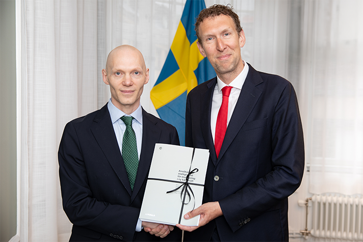 Two men in suits looking facing the camera, holding papers and shaking hands. Swedish flag in background. Indoors.