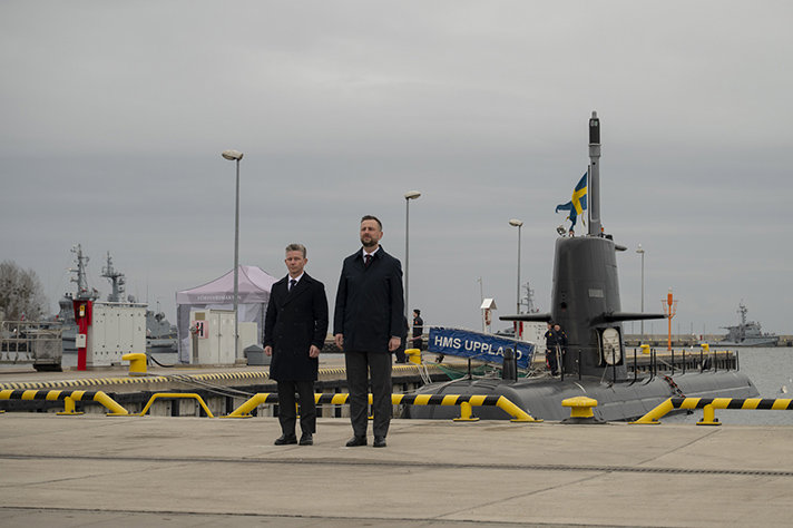 Pål Jonson and Władysław Kosiniak-Kamysz stands at attention by a dock in Gdynia. It's a cloudy day and in the background the swedish submarine HMS Uppland is docked.