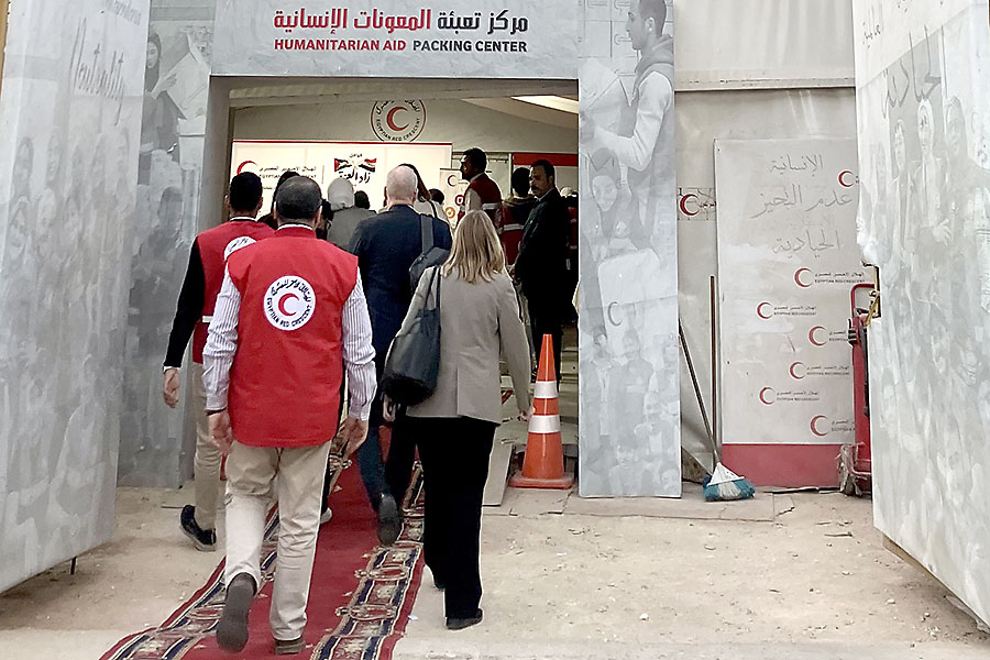 People enter a humanitarian aid‑packing centre, guided by staff in red vests bearing the Red Crescent emblem. The entrance is decorated with banners and photographs, and a red carpet covers the ground.
