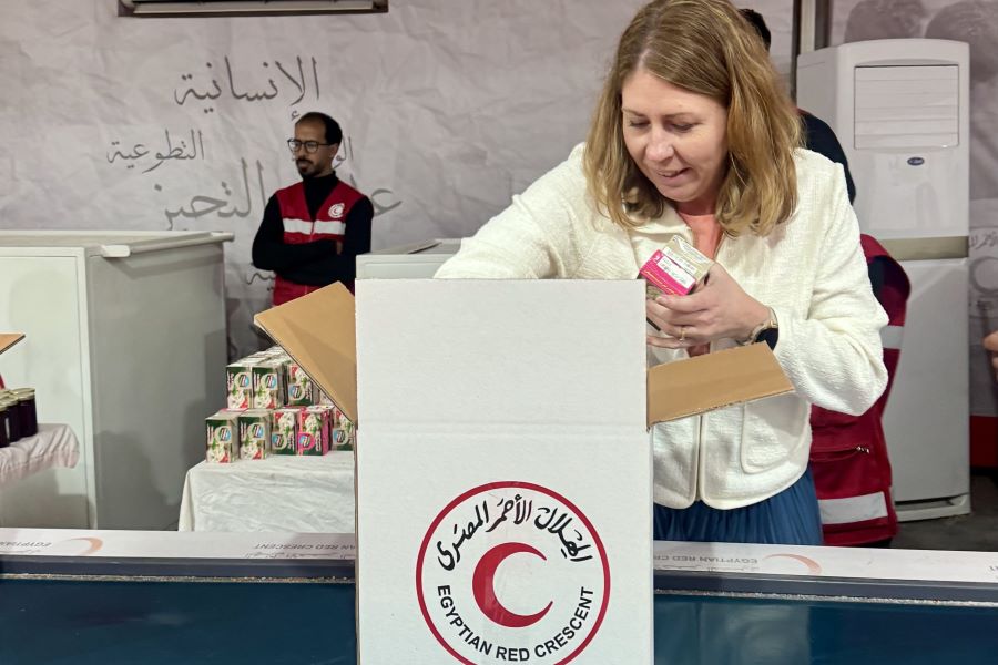 Minister for Health Care Elisabet Lann packs humanitarian supplies into a box labelled ‘Egyptian Red Crescent’. In the background, staff in red vests and stacks of relief items can be seen.