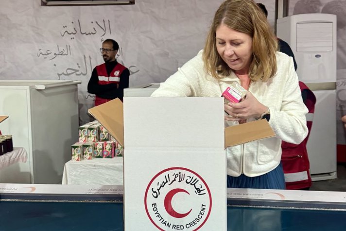 Minister for Health Care Elisabet Lann packs humanitarian supplies into a box labelled ‘Egyptian Red Crescent’. In the background, staff in red vests and stacks of relief items can be seen.